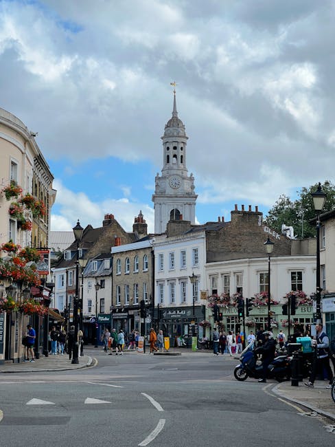 A busy street scene in Greenwich featuring classic terraced shops and pedestrians on the pavement, with a white historical clock tower and spire rising prominently in the background under a partly clo