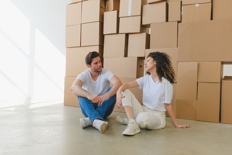A man and a woman are engaged in a home relocation process indoors, surrounded by numerous cardboard moving boxes of various sizes, some sealed and others open showing packing materials. The man, wear