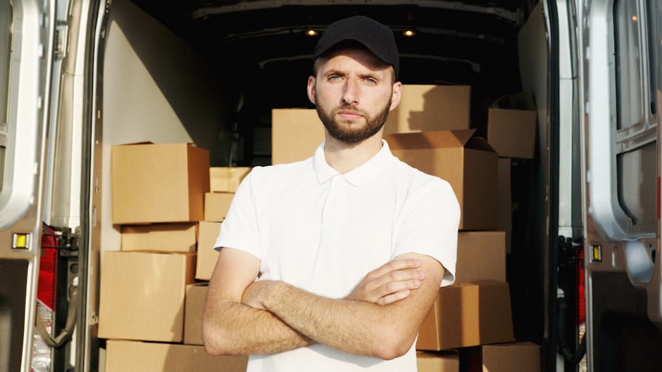 A young man with a beard, wearing a black cap and a white polo shirt, stands inside the open rear doors of a moving van filled with neatly stacked cardboard boxes of various sizes wrapped in plastic a