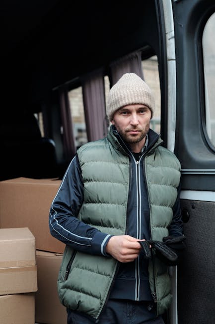 A young man with a beard, wearing a black cap and a white polo shirt, stands inside the open rear doors of a moving van filled with neatly stacked cardboard boxes of various sizes wrapped in plastic a
